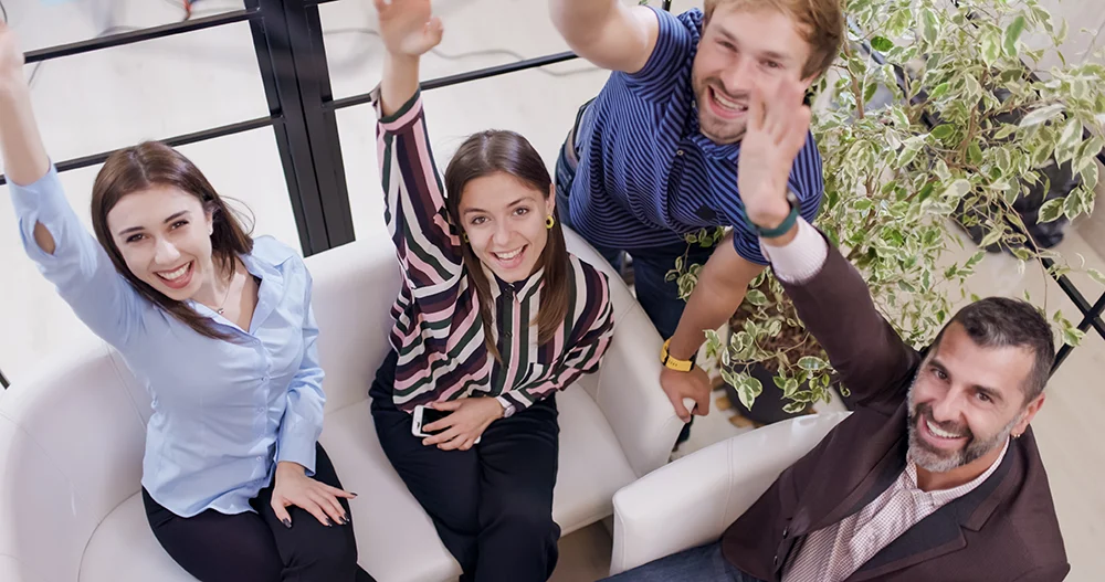 4 people sit together in a bright office area. They are smiling and waving at the camera.