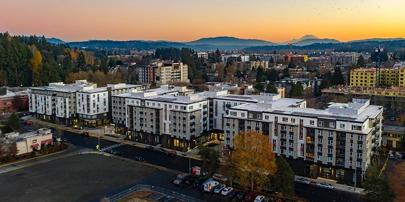 Aerial view of a large commercial residential complex surrounded by streets, trees, and nearby buildings at sunset, with mountains in the distance.